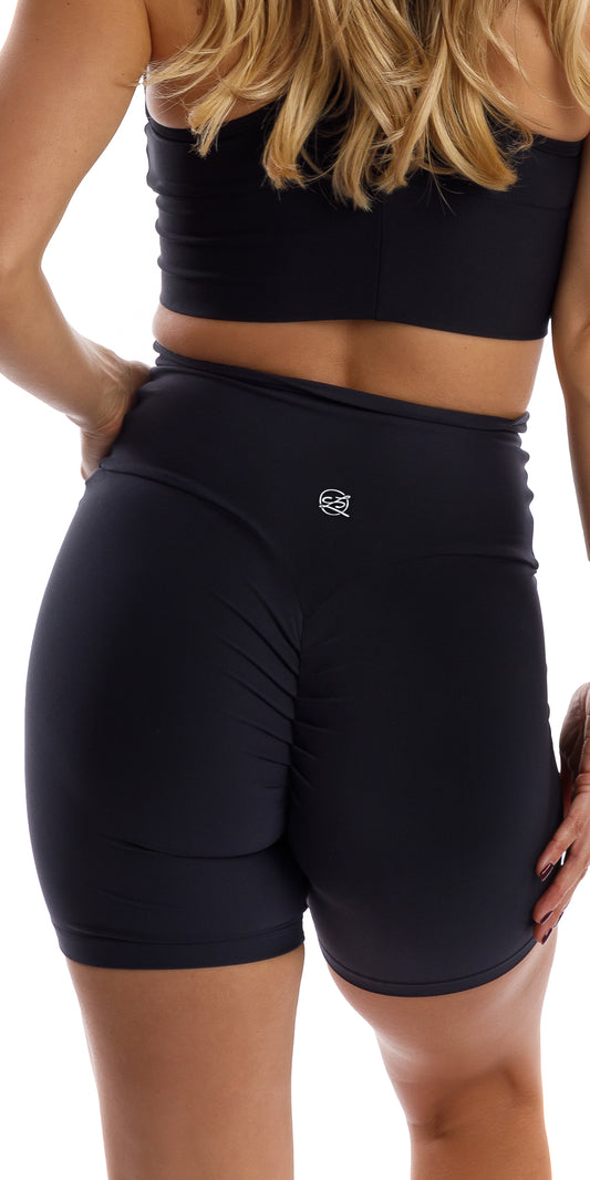 Back view of girl wearing black Midnight Eco Scrunch Bum Midi Shorts putting one hand on waist