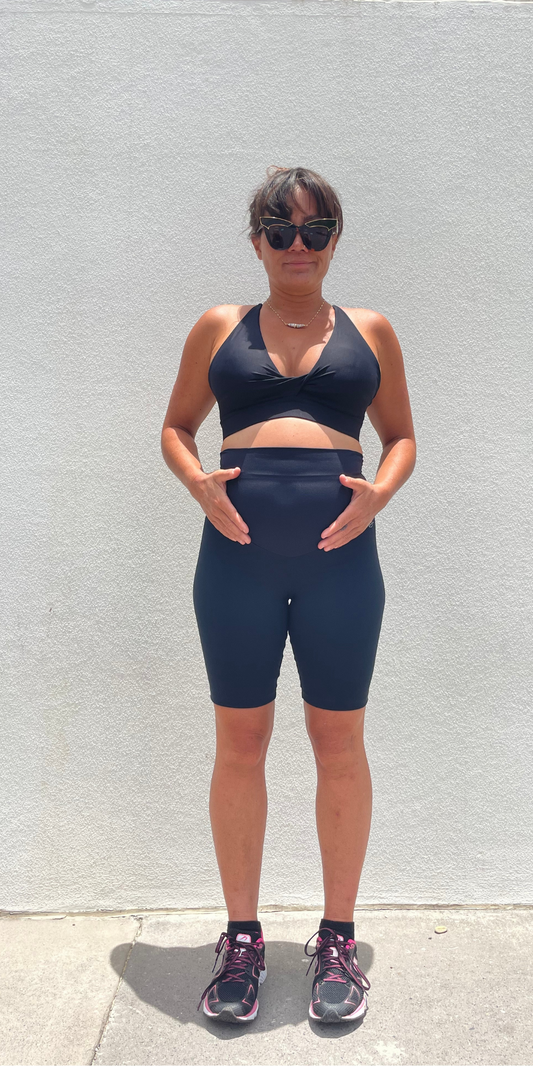 Full body front view of woman in black Midnight Maternity Biker Shorts and matching bra touching her baby bump