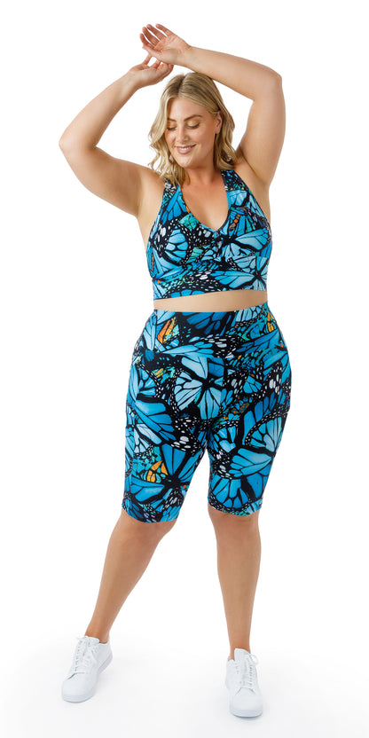 Full body front view of girl in blue animal print JH Butterfly Biker Shorts with Pockets and matching sports bra lifting both arms all the way to the top of her head