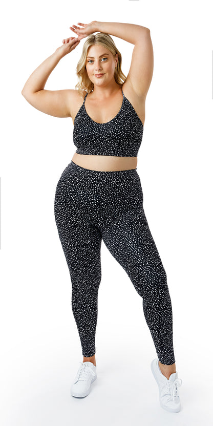 Full body front view of lady in black Star Dust Momentum Bra and matching leggings putting left foot out lifting its heel while raising both arms