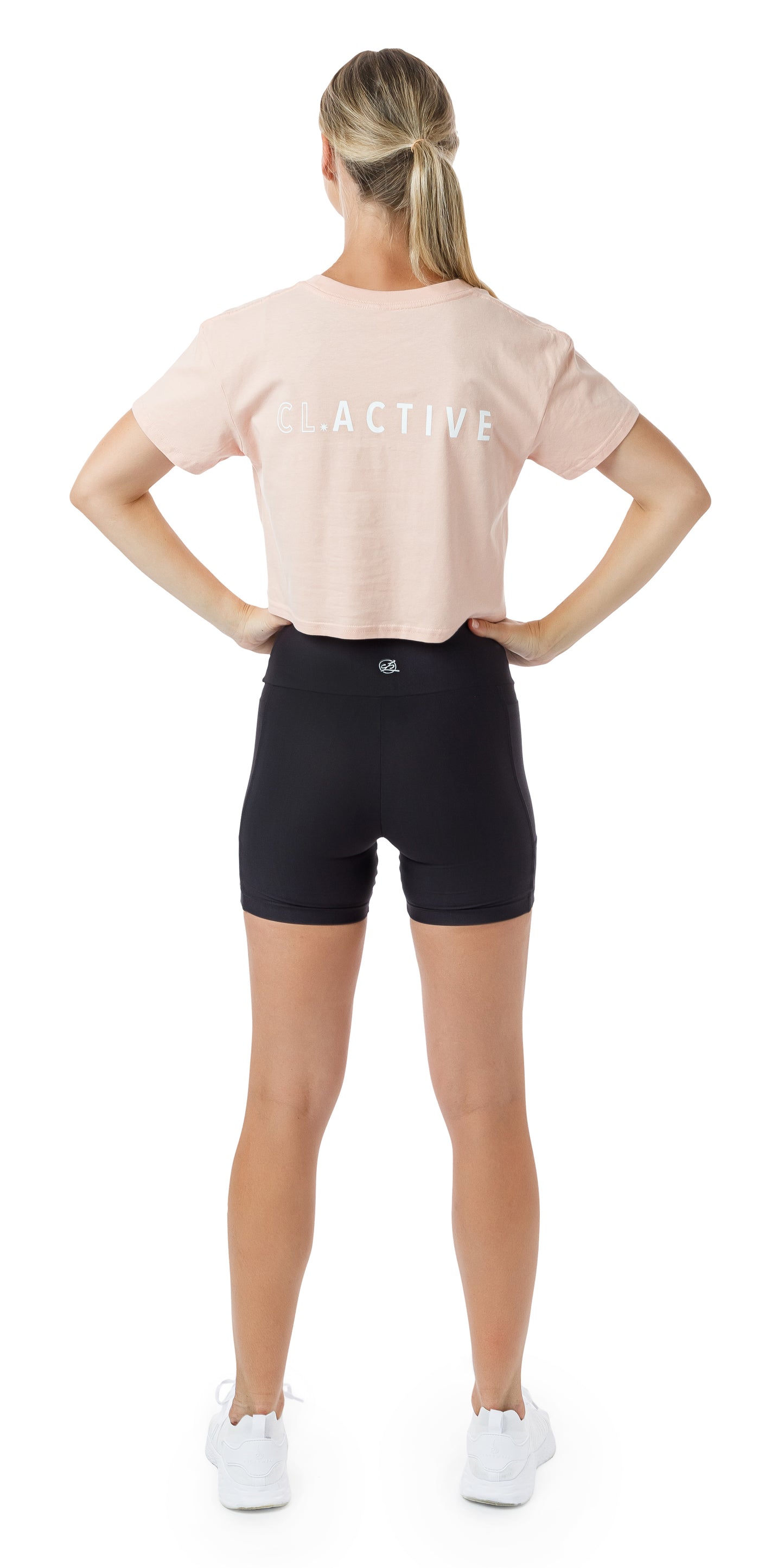 Full body rear view of girl in Pink CL Active Crop Tee and black biker shorts putting both hands on waist