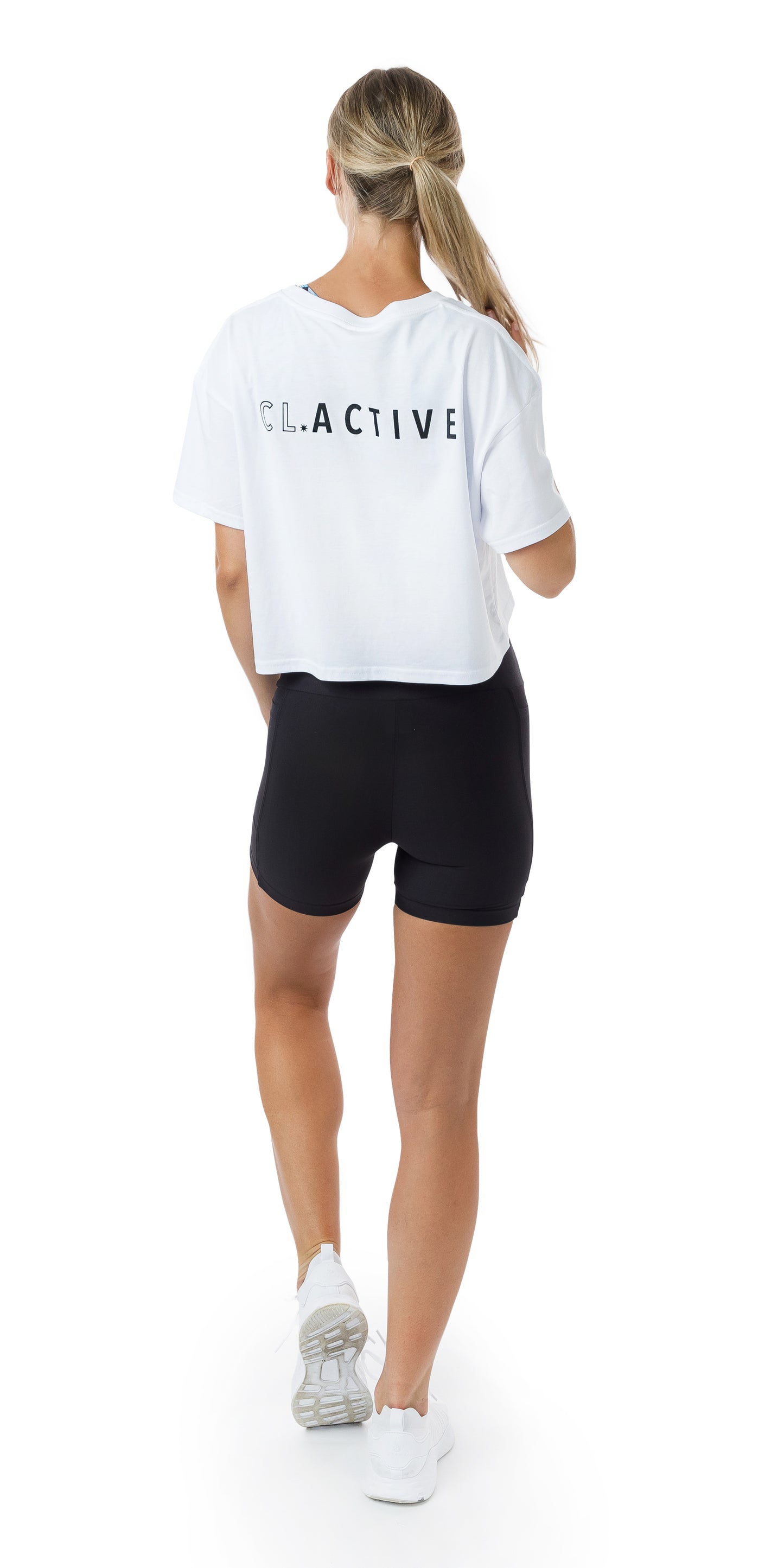 Full body back view of lady in White CL Active Crop Tee and black short lifting left heel and holding her ponytail