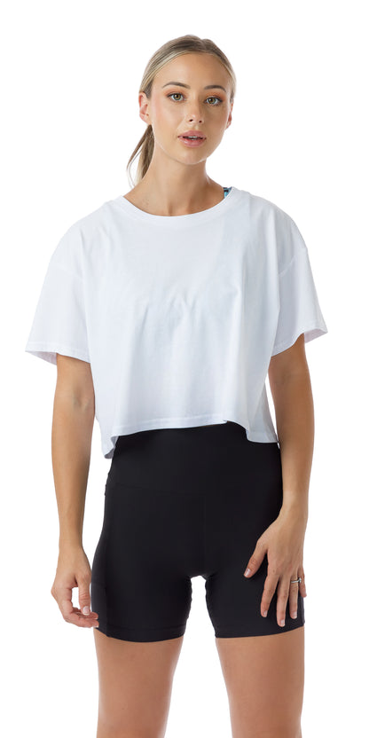 Front view of lady in White CL Active Crop Tee and black shorts standing and putting left hand on lap