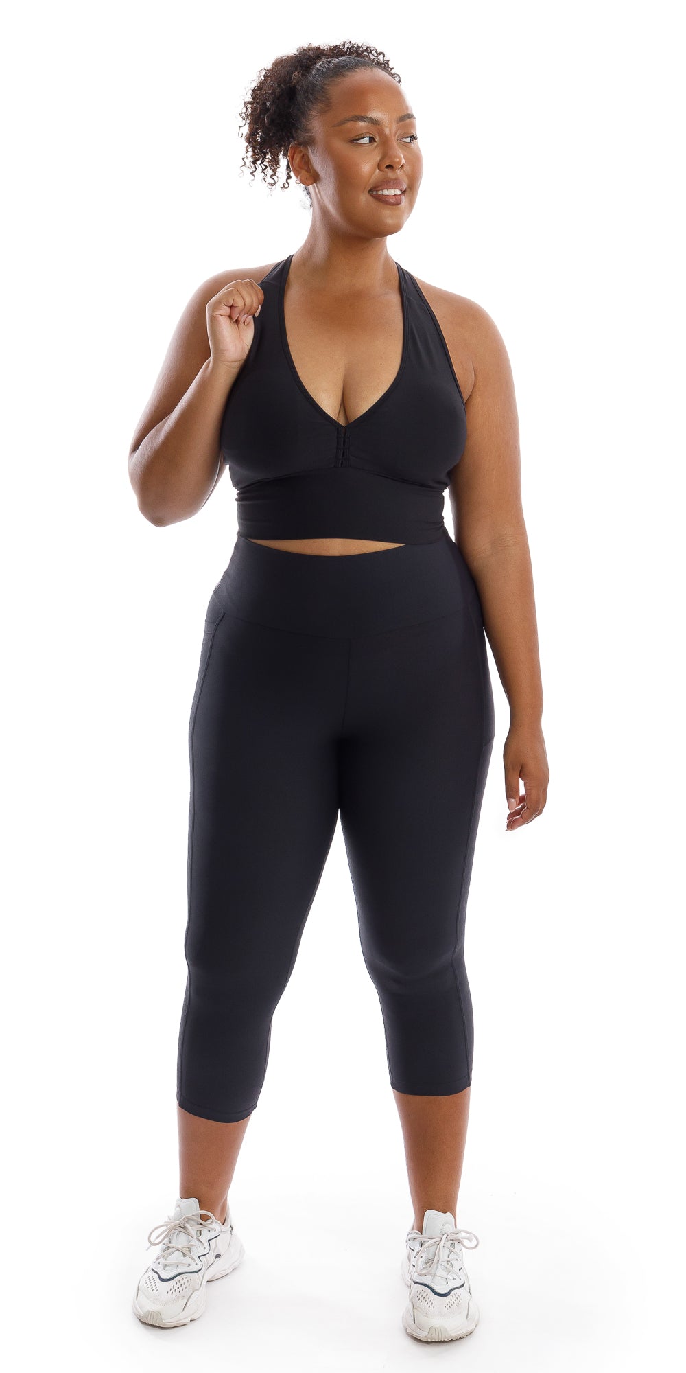 Full body front view of girl in black Midnight Body Luxe Capri Leggings with Pockets and matching bra bringing one arm close to her shoulder and looking to her left