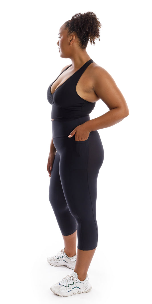Full body side view of girl in black Midnight Body Luxe Capri Leggings with Pockets and matching bra putting one hand in pocket