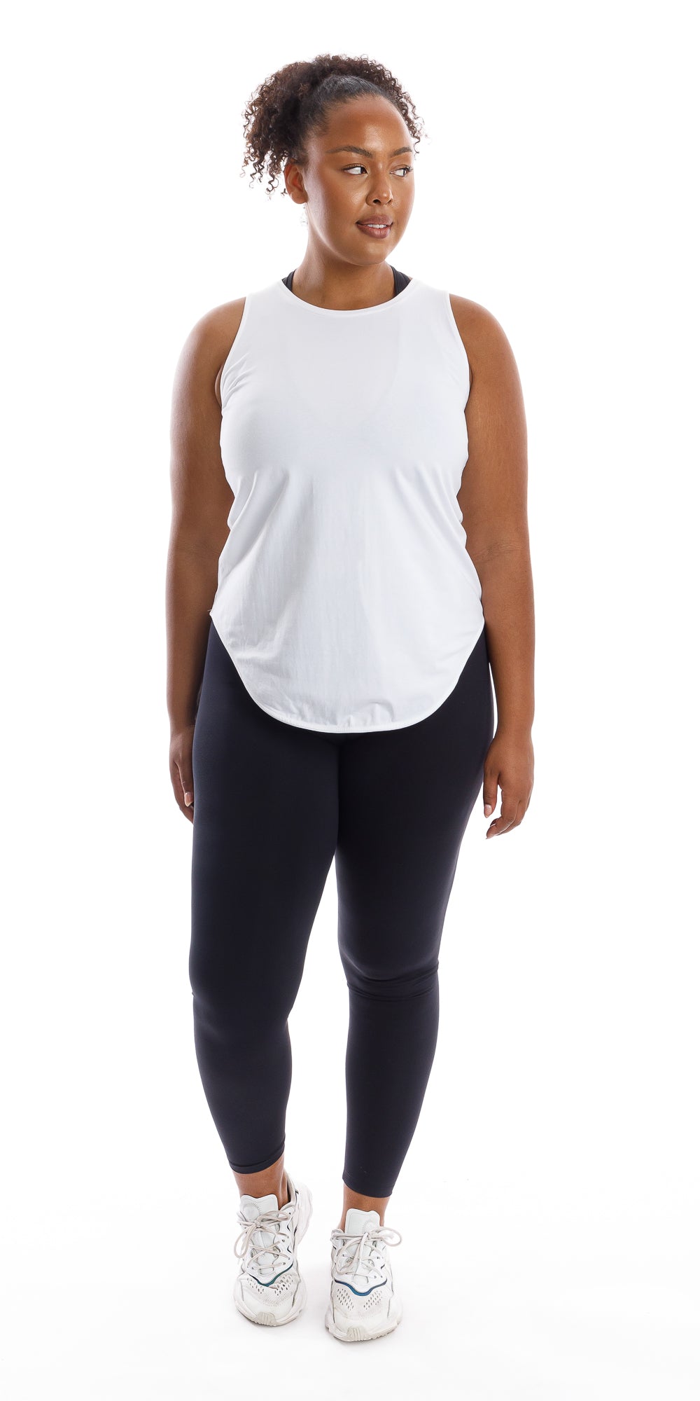 Full body front view of lady in sleeveless White Palm Beach Tank and black leggings lifting right heel and looking aside