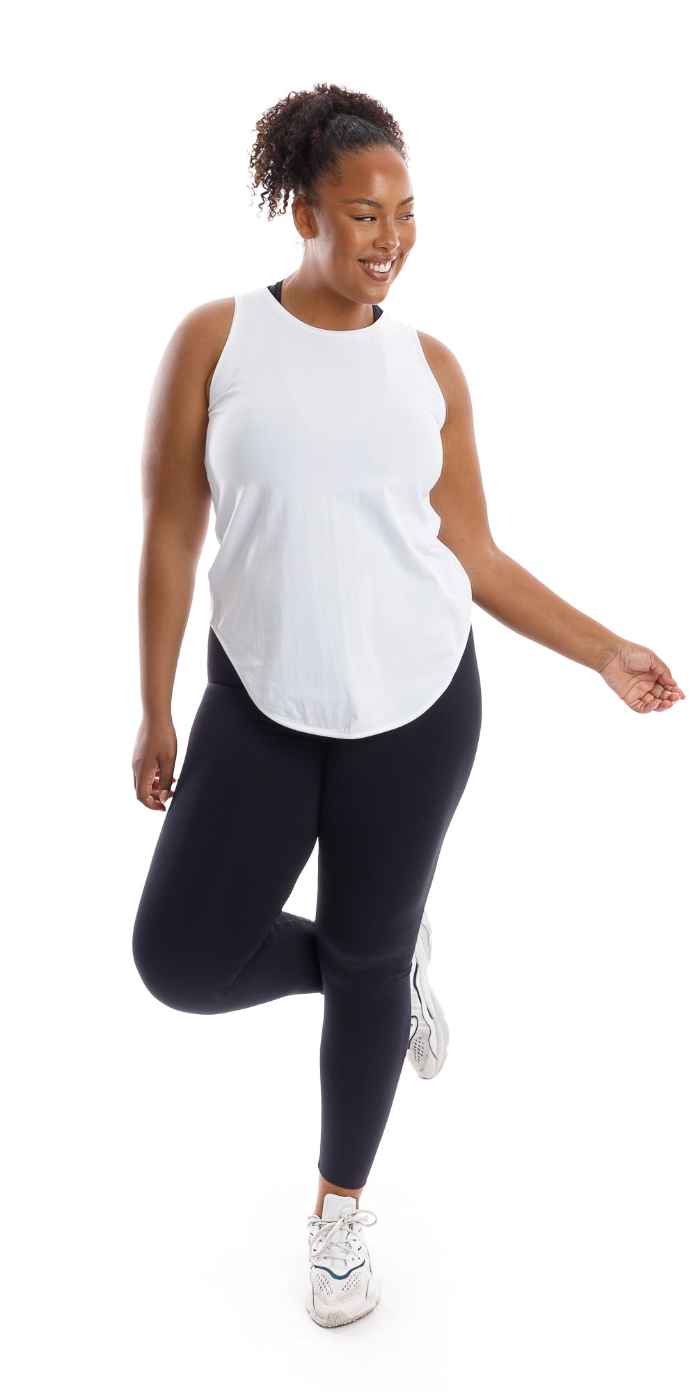 Full body front view of lady in sleeveless White Palm Beach Tank and black leggings standing on left leg lifting right leg crossing it behind while swaying arms and smiling