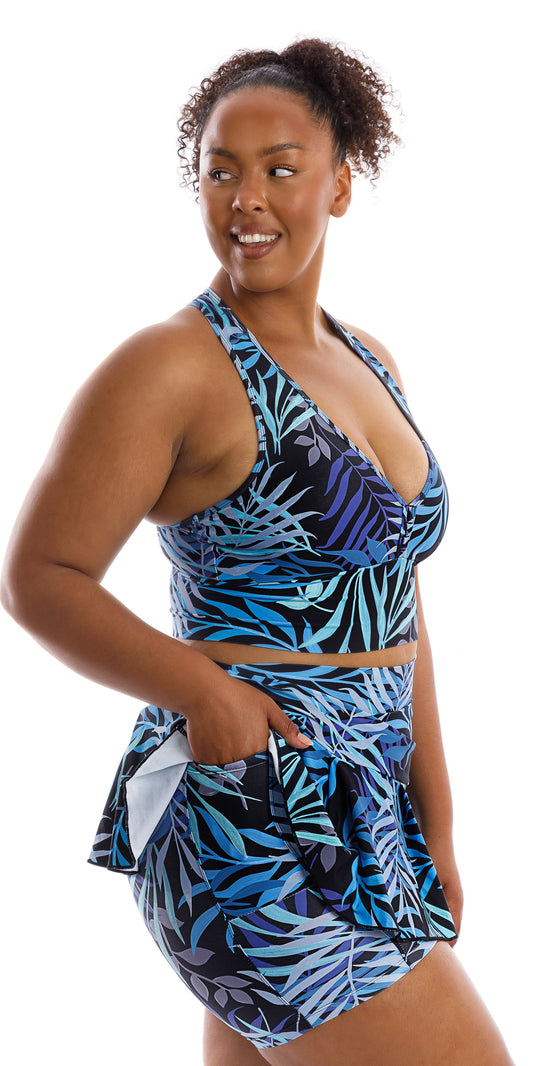 Angled side view of lady in black and blue Tropical Palm Skort with Pockets and matching bra putting right hand in the pocket and looking aside while smiling