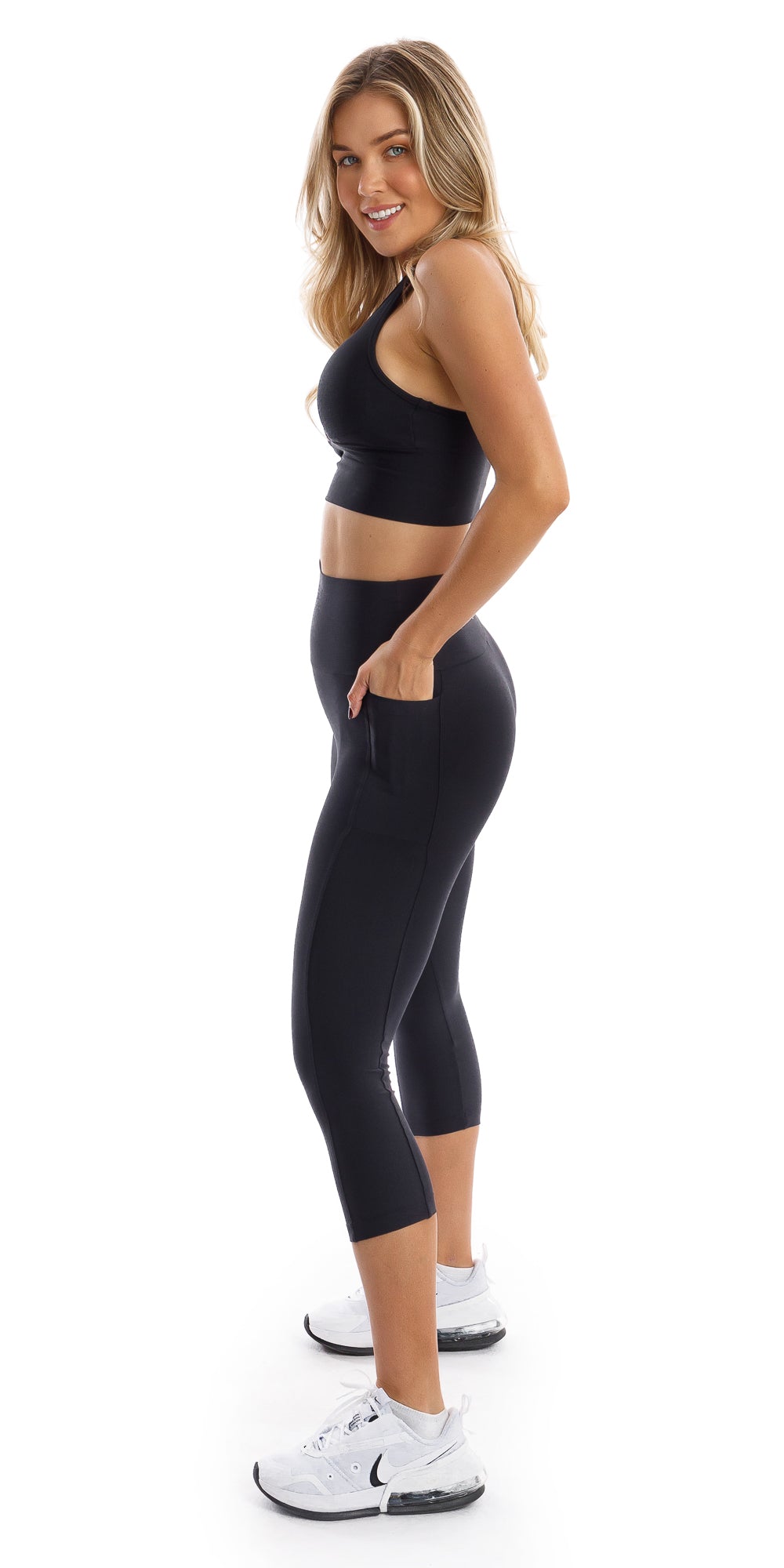 Full body side view of girl wearing black Midnight Eco Capri Leggings with Pockets and matching bra putting hands in the pockets