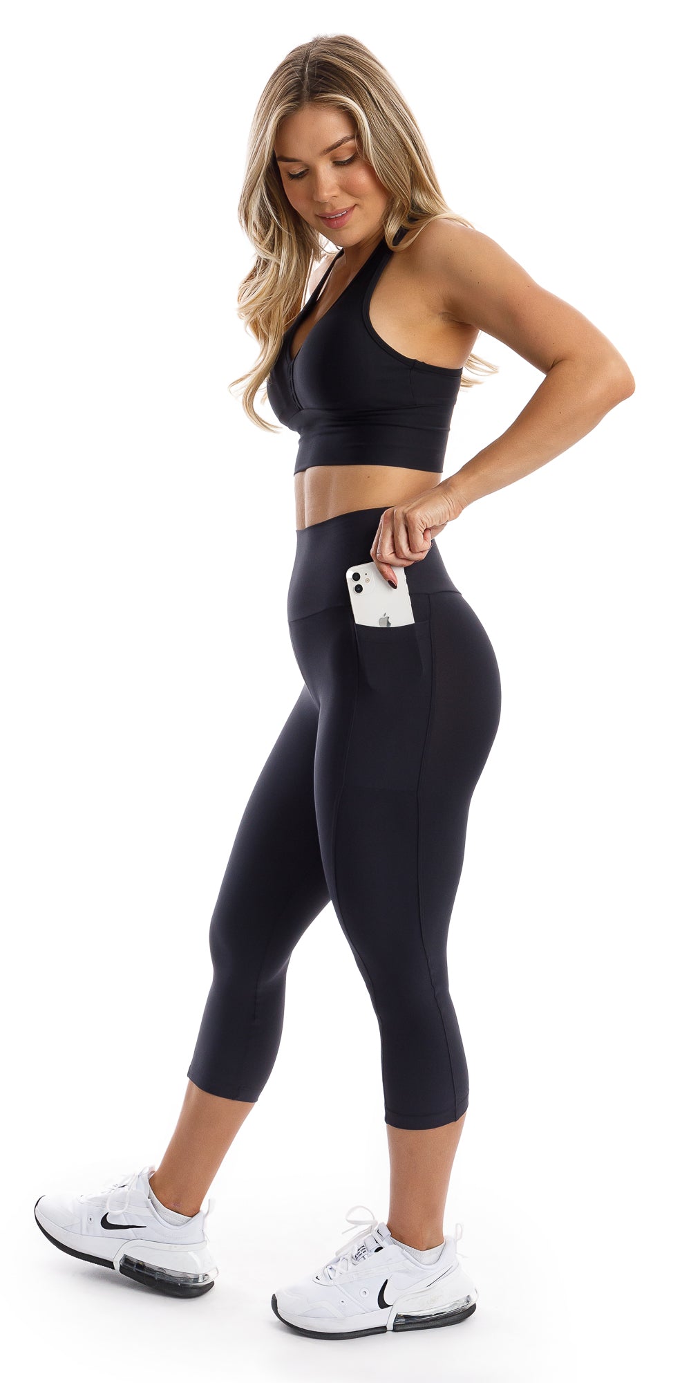 Full body side view of girl wearing black Midnight Eco Capri Leggings with Pockets and matching bra putting phone in the pocket