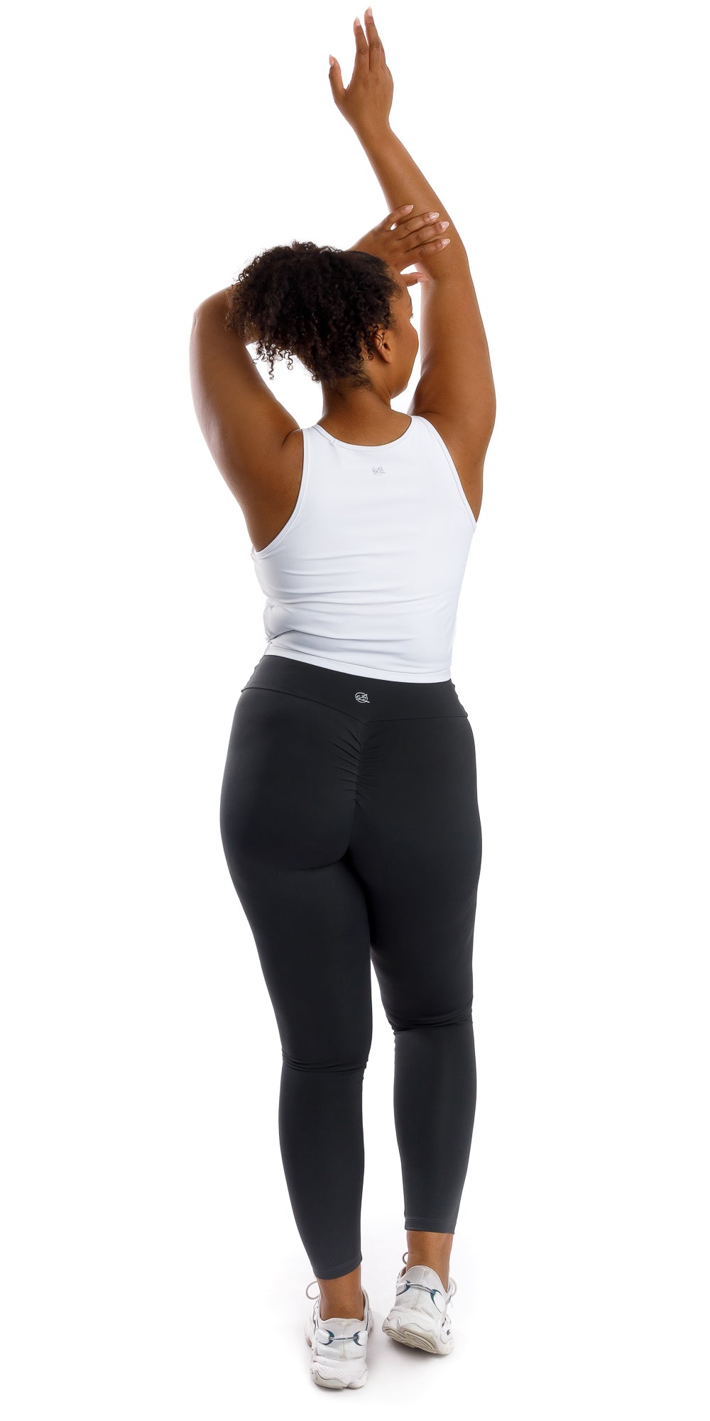 Full body back view of lady in sleeveless White Crop Top and black leggings lifting right heel and raising both arms with one all the way up while folding the other
