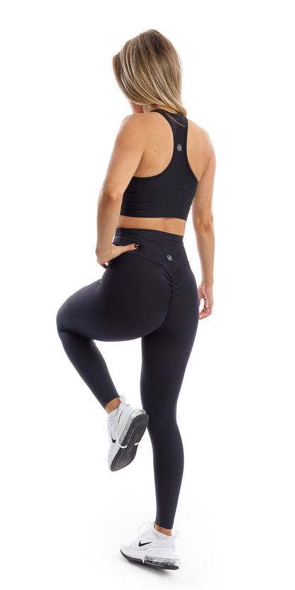 Angled side to rear view of girl wearing black Midnight Eco Scrunch Bum Leggings and matching bra lifting one leg and putting one hand on waist