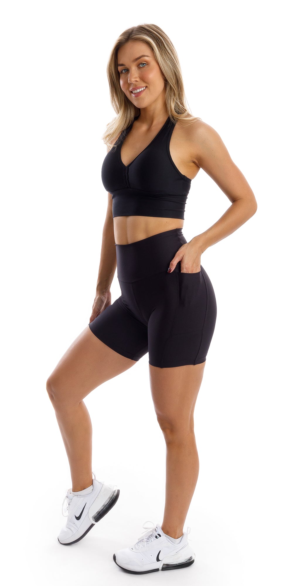 Full body side view of girl in black Midnight Eco Midi Shorts with Pockets and matching bra putting one hand in pocket