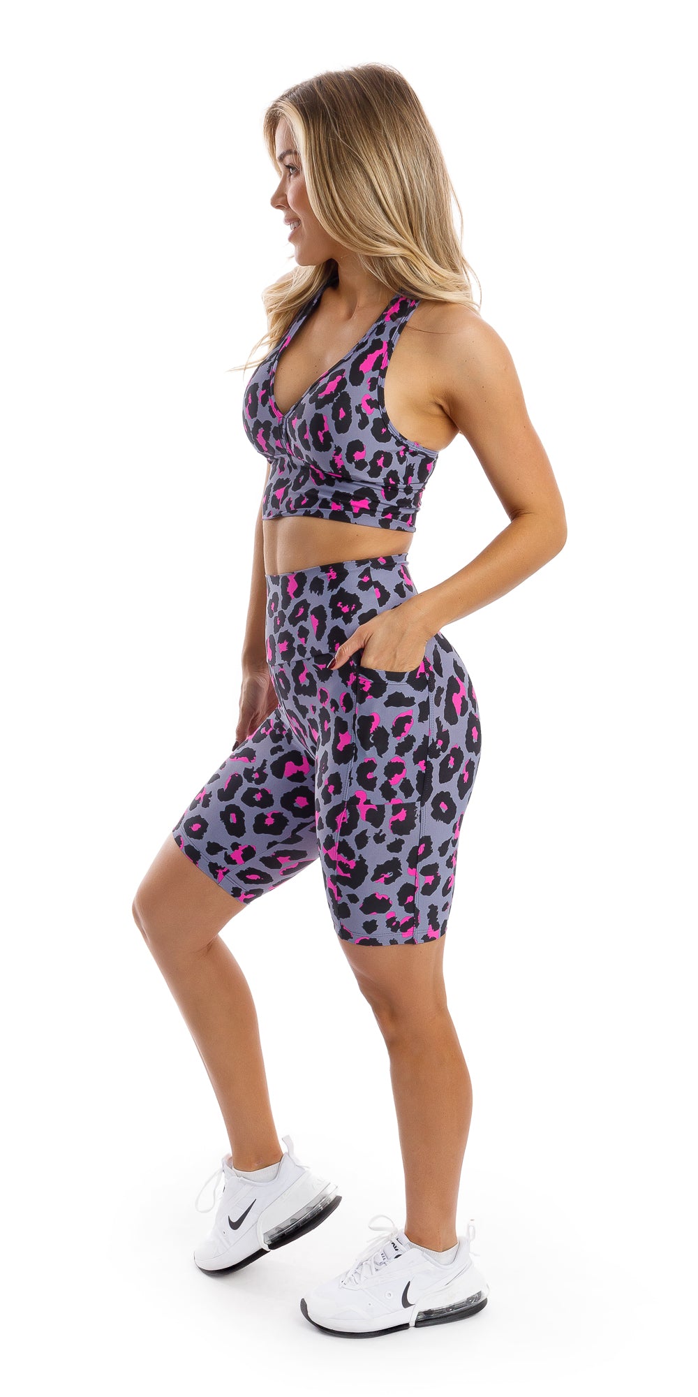Full body side view of girl in animal print Pink Leopard Eco Biker Shorts with Pockets and matching bra putting bent knee forward and one hand in pocket