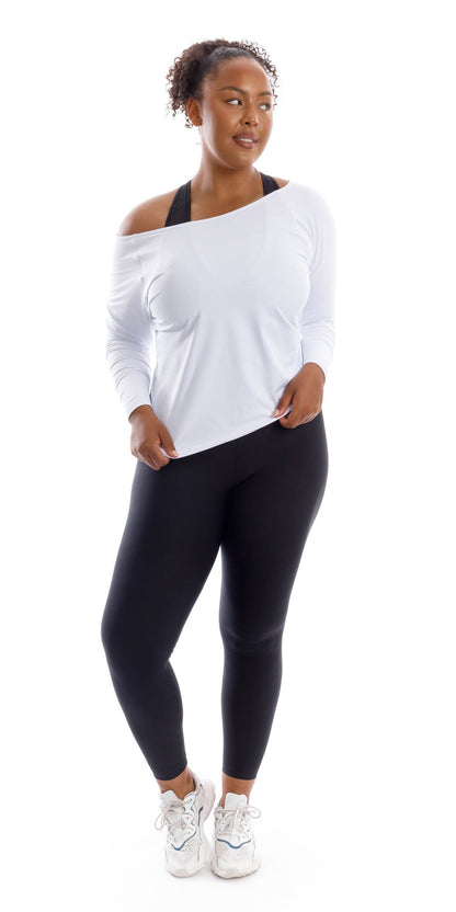 Full body front view of lady in White Off The Shoulder Long Sleeve Tee and black leggings lifting right heel and touching the end of the shirt with both hands while looking aside