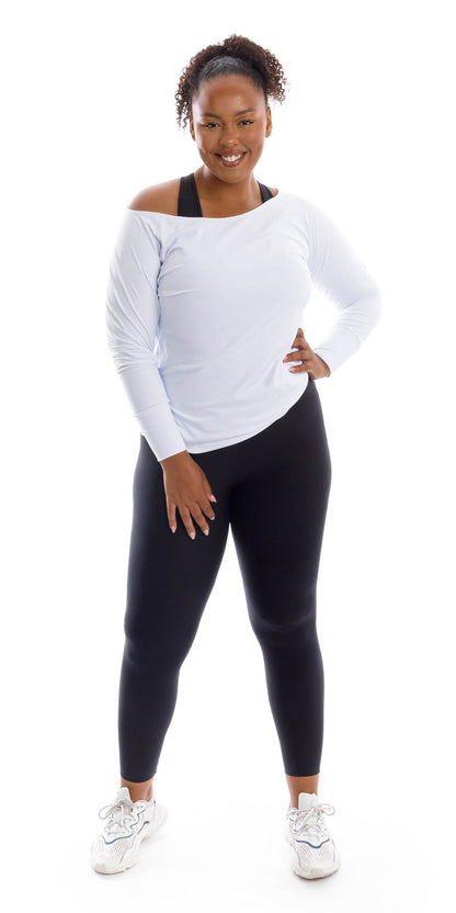 Full body front view of lady in White Off The Shoulder Long Sleeve Tee and black leggings standing with feet apart and putting left hand on waist while smiling