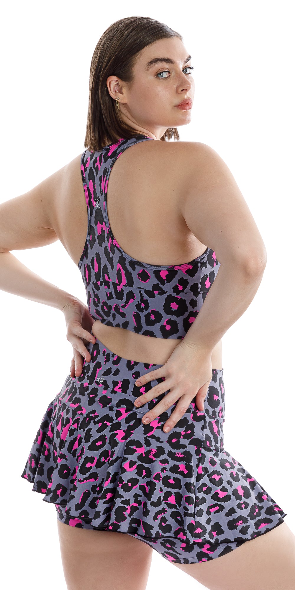 Angled side view of girl in animal print Pink Leopard Eco Racer Back Bra and matching skort looking over her right shoulder and putting both hands on waist
