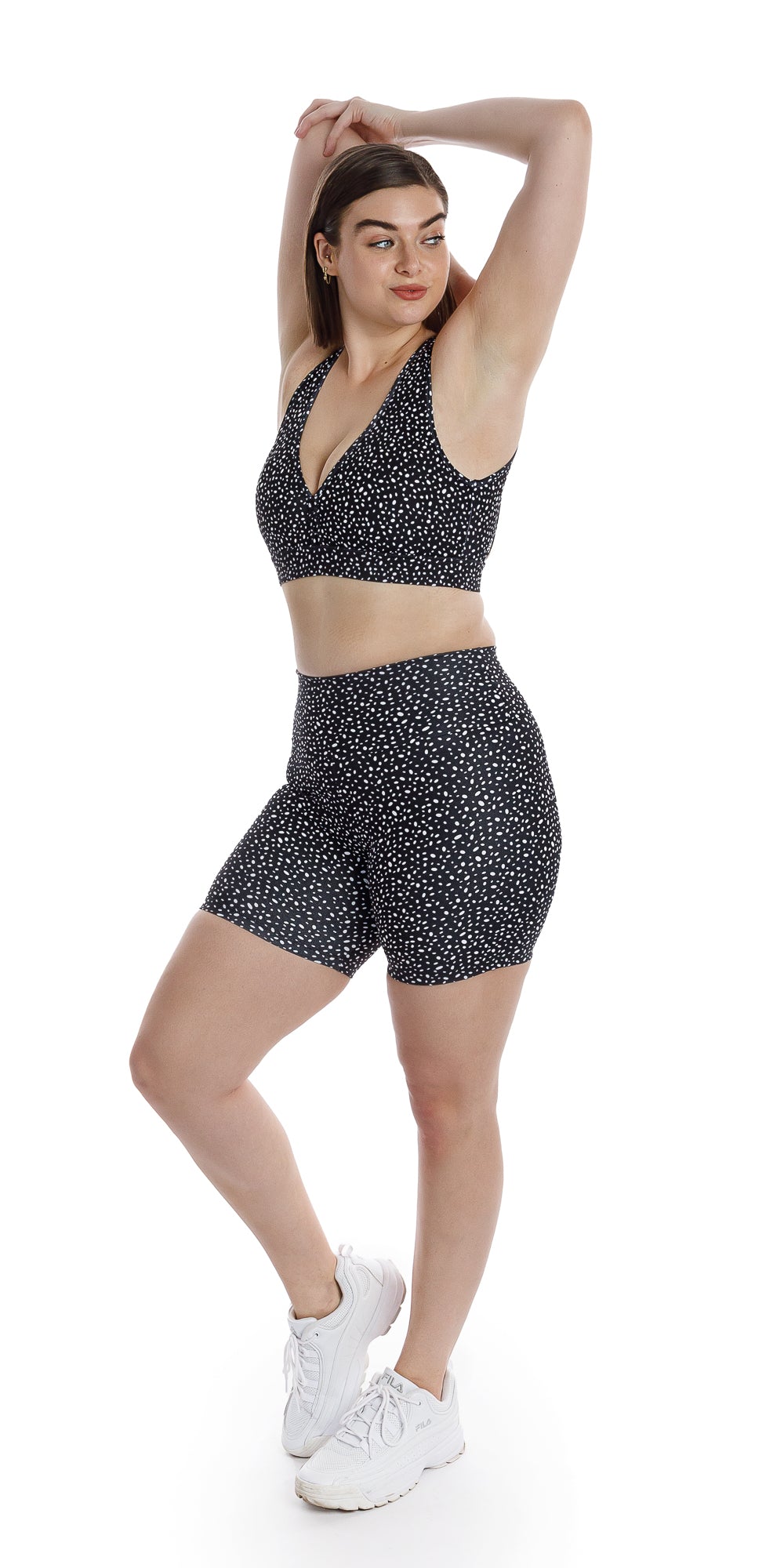 Full body angled front view of lady in black Star Dust Rhythm Bra and matching bottoms looking aside and lifting right heel while putting both arms behind her head