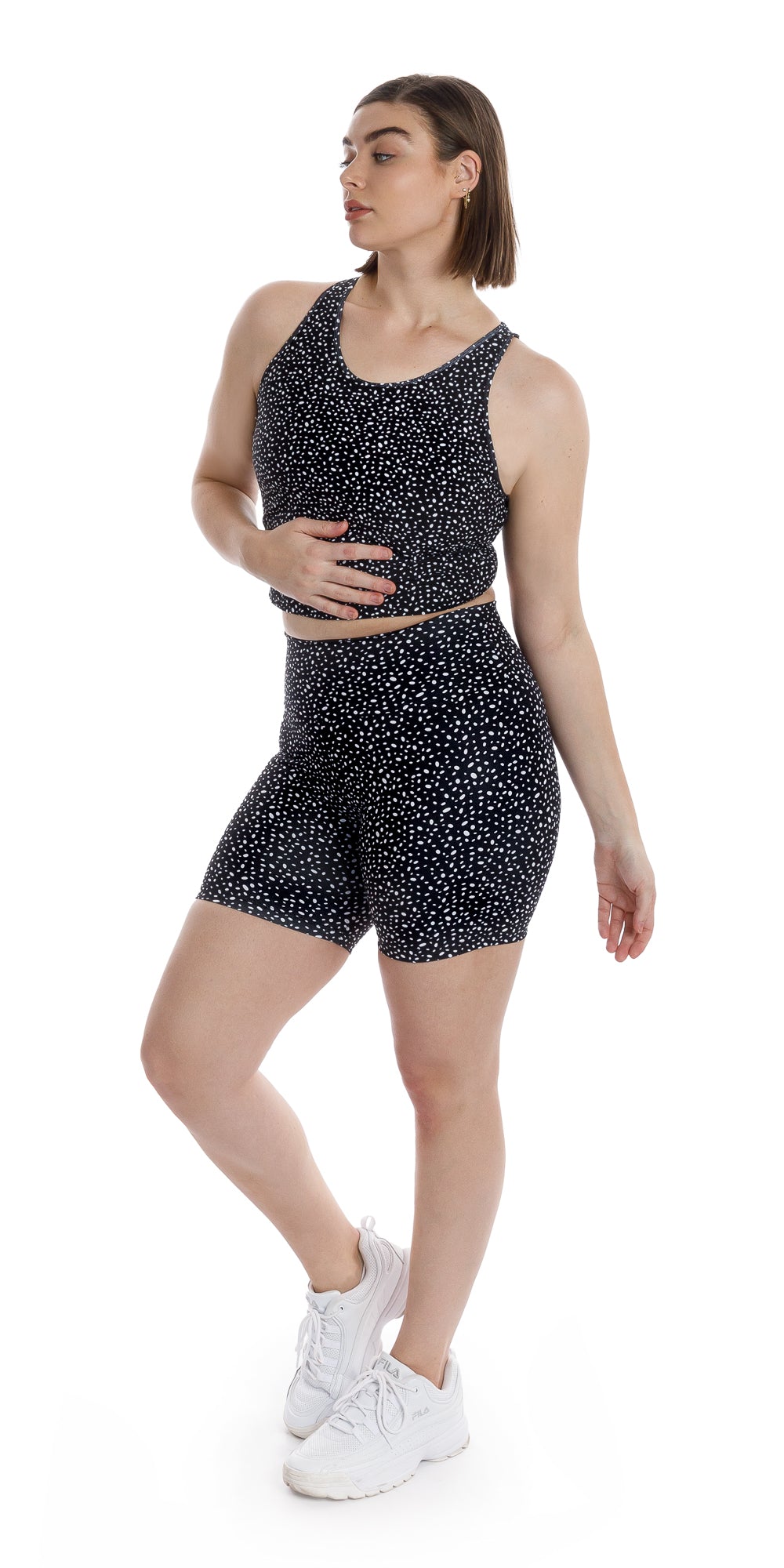 Front view of girl in black Star Dust Body Luxe Midi Shorts and matching top looking aside while lifting right heel and putting right hand on stomach