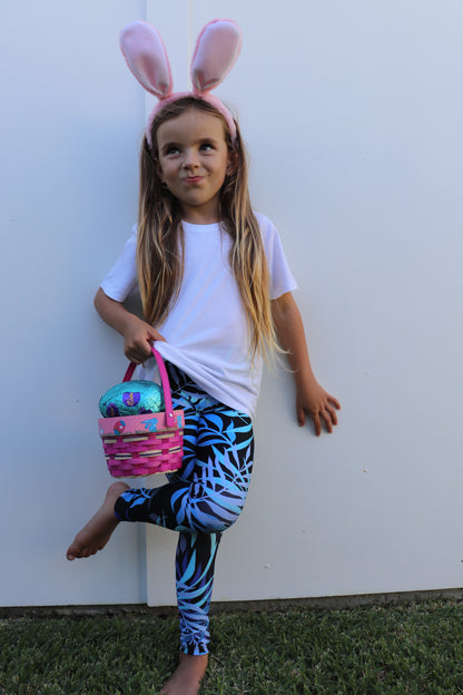 Full body front view of girl in printed black Tropical Palm Mini Me Leggings and white shirt crossing left leg over right leg while holding a pink basket of toys