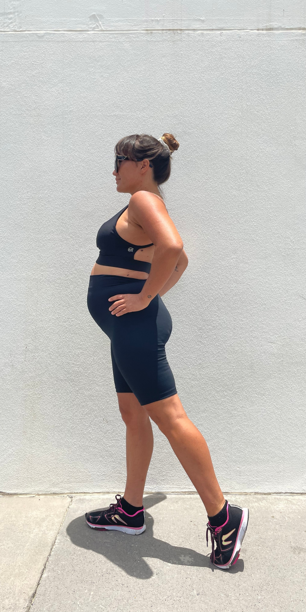 Full body side view of pregnant woman in black Midnight Maternity Biker Shorts and matching bra putting both hands on waist